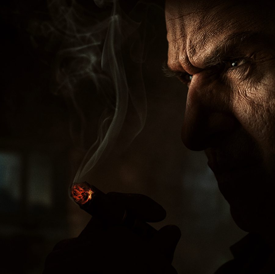In a dark room, villain Oz Cobb (Colin Farrell) glares menacingly into the distance while smoking a cigarette, in a scene from The Penguin. 